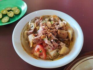 Bowl of soto ayam with shredded chicken, tomato, crispy shallots, noodles, and clear savory broth, served with lime on the side and steamed rice.