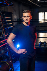 Confident man poses in fitness studio with equipment nearby. Man stands confidently in a fitness studio, surrounded by gym equipment and vibrant lighting.