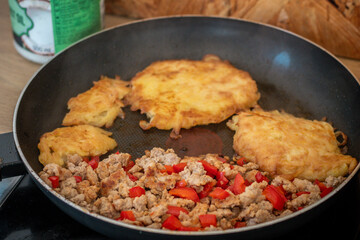 A frying pan on a stovetop with crispy golden-brown potato pancakes (latkes or hash browns) cooking alongside seasoned ground meat mixed with chopped red bell peppers.