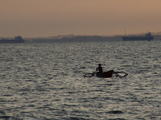 Fototapeta premium Silhouette of a traditional fisherman in a small outrigger boat on the open sea, with large cargo ships and a coastal skyline in the distant background at sunset.