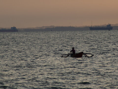 Silhouette of a traditional fisherman in a small outrigger boat on the open sea, with large cargo ships and a coastal skyline in the distant background at sunset. - Powered by Adobe