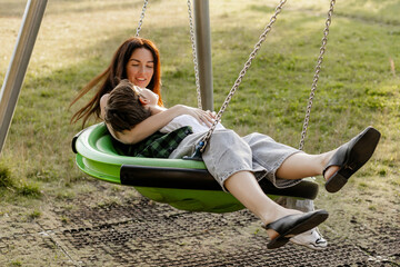 Mother and son sitting together on green swing at playground, enjoying tender family moment in nature on sunny day