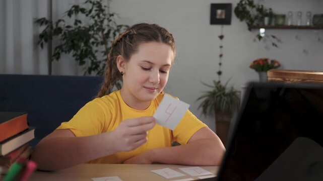 Studying with flashcard and laptop student girl child in yellow shirt at desk sorting study card while smile and focusing on homework and learning with book pen plant nearby calm concentration