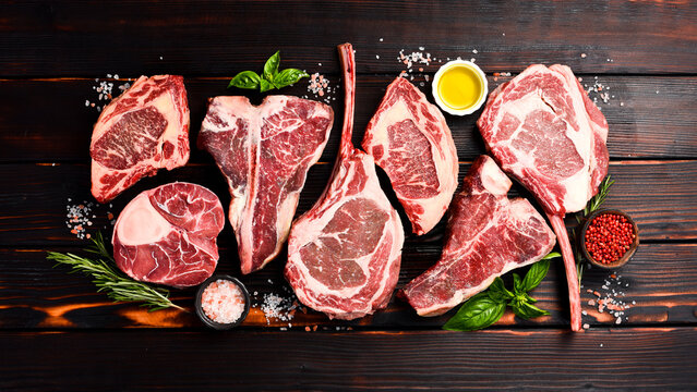Variety of raw beef meat steaks for grilling with seasoning: Tomahawk steak, Ribeye, Ossobuco and T-bone steak, on a wooden brown background. - Powered by Adobe