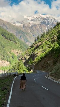 4K Vertical shot of Indian woman walk on asphalt road and snow mountains in background at Pangi Valley, Chamba, India. Monsoon clouds in sky creating scenic view. Tourist exploring Himalayas.