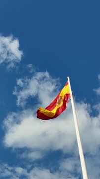 The national flag of spain fluttering on a flagpole against a beautiful blue sky with clouds