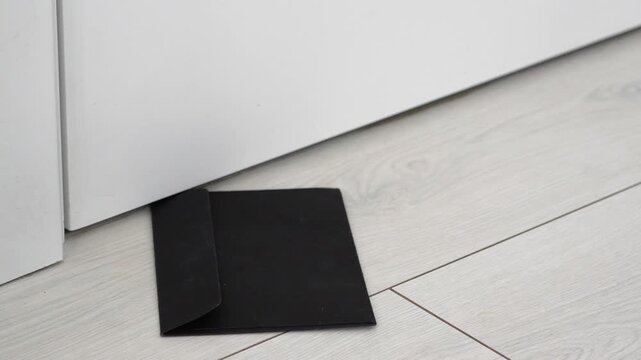 The postman slips an envelope containing a letter under the door of a house.
Letter delivery with a message.
Black envelope with a handwritten letter.
Delivery, letter, information, under, door