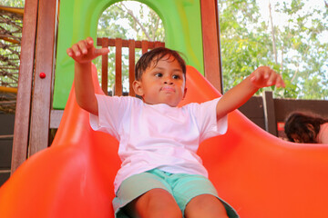 A young Asian boy with a determined expression sits on a red playground slide with his hands...
