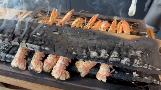 Des langoustines enti&egrave;res coinc&eacute;es entre deux planches de bois brul&eacute;es, en train de fumer et de finir lentement leur cuisson au feu de bois.