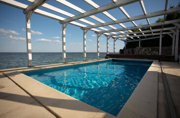 Outdoor swimming pool with clear blue water under a wooden pergola by the sea. Scenic view of the ocean horizon and relaxing summer atmosphere.