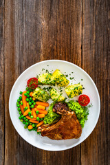 Baked veal cutlet with bone, boiled potatoes, mini carrots and green peas on wooden table