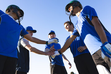 Teenage baseball players in blue jerseys and helmets huddling on sunny field stacking gloves