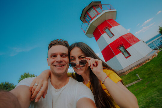 Smiling couple taking a cheerful selfie in front of a red and white striped lighthouse on a sunny day, symbolizing travel, love, happiness, and adventure.