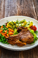 Baked veal cutlet with bone, boiled potatoes, mini carrots and green peas on wooden table