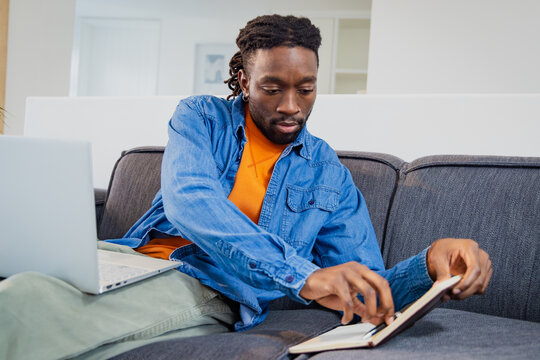 African American man flipping through notebook on grey sofa at home by silver laptop