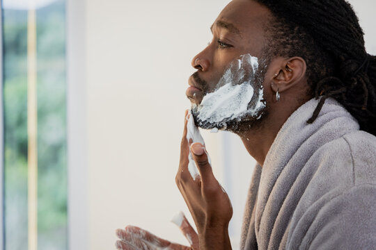 African American man wearing gray bathrobe applying shaving foam at bathroom mirror by window frame