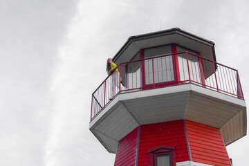 Smiling young woman in yellow shirt leans on the balcony of a red and white lighthouse, enjoying the view on a clear day with a bright blue sky and scattered clouds.