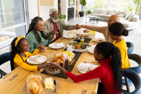 Multigenerational family joining hands and saying grace at home dining table with plates and turkey