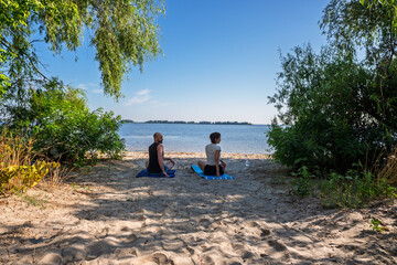 A serene couple meditates on a sandy beach by a tranquil lake under a clear blue sky, embraced by nature's peace. Ideal for themes of wellness, mindfulness, and outdoor relaxation.