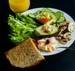 Side Shot of Vegetarian Breakfast with Avocado, Mushrooms, Green Lettuce, Soft Cheese with Red Pepper, Cucumbers, Tomatoes, Seeded Multigrain Bread and Orange Mango Fresh Juice