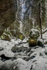 The image depicts the picturesque landscape of the Czech Switzerland National Park. The picture shows a snow-covered stream in a forest.Czech Republic.
