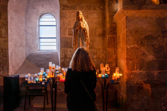 Silent devotion of a woman with her back turned praying before the image of the Virgin Mary with votive candles inside a medieval stone church.