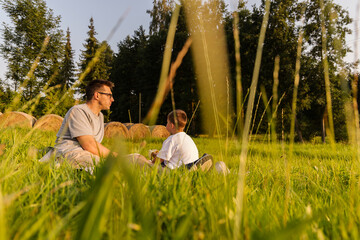 A father and son relax on a grassy field, sharing stories and laughter. They enjoy the warmth of the sun and the beauty of nature surrounding them