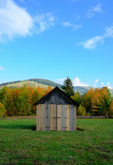 A small weathered shed sits in a green field, surrounded by autumn trees. Quiet countryside scene with a bright sky, capturing rustic charm and peaceful rural life.
