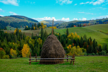 A large haystack enclosed by a rustic wooden fence sits in a green field under a bright blue sky. Autumnal forests and rolling hills create a peaceful rural landscape.