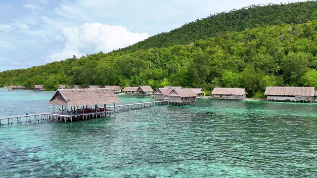 Aerial view of huts with thatched roofs over the turquoise water with lush green forest in the background, Kri Island Resort, Raja Ampat, West Papua, Indonesia.