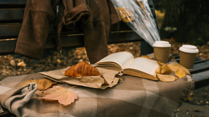 Autumn picnic scene with coffee, croissant, and a book on a park bench.