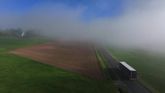 Aerial view of a lone truck making its way down a road amidst a dense fog, bordered by green fields, Eichenzell, Hessen, Germany.