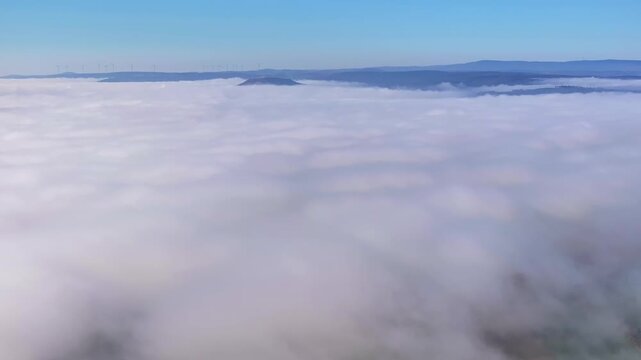 Aerial view of rolling white clouds blanketing hills and distant mountains under a clear blue sky, creating a serene and ethereal landscape, Eichenzell, Hessen, Germany.
