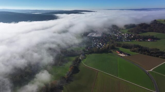 Aerial view of houses nestled under a blanket of fog, contrasting with the bright green fields, Eichenzell, Hessen, Germany.