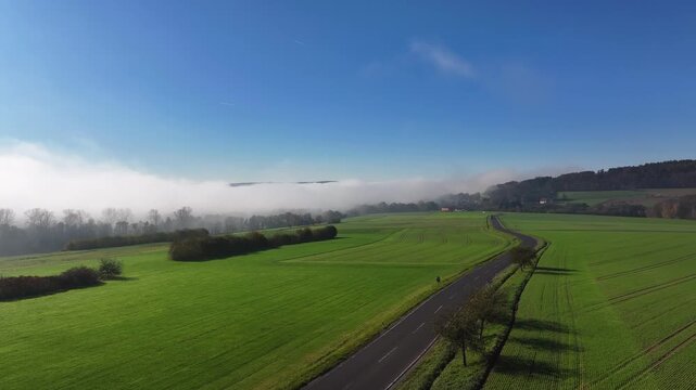 Aerial view of lush green fields contrasting with a thick blanket of fog partially obscuring distant hills and a winding road, Eichenzell, Hessen, Germany.