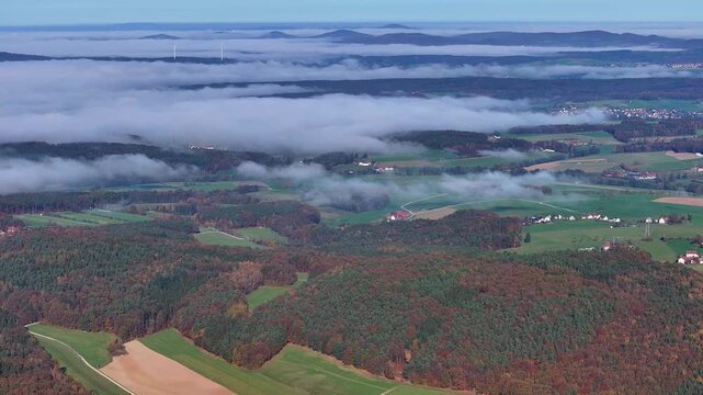 Aerial view of a dense forest meeting a sea of ethereal clouds, with distant hills peeking through the mist, Eichenzell, Hessen, Germany.