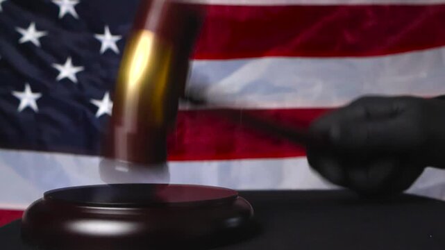Close up view of a gloved judge justice hand hitting a court gaol wooden and bronze hammer on a black table while an American colorful United States flag is in the background. Slow motion movement.
