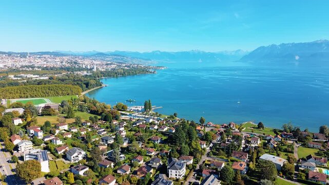 Aerial view of Lake Geneva's shimmering expanse, bordered by the picturesque town, lush greenery, and distant mountains, Saint-Sulpice, Vaud, Switzerland.