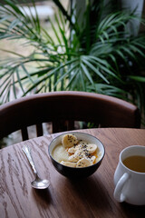 Sweet polenta porridge with caramel, banana and black sesame. Side view, wooden table, cappuccino, tropical leaves, milk, wheat, white, semolina, pudding.