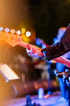 A musician, wearing a watch, plays a bass guitar on stage during a live performance, with the instrument's headstock and tuning pegs visible, illuminated by colorful stage lights and a music stand nea