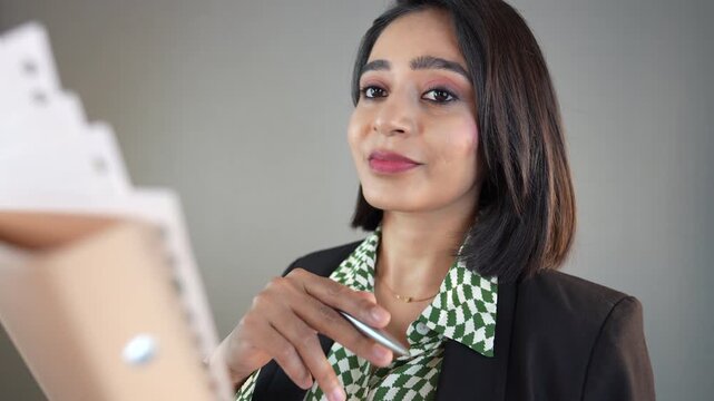 Closeup portrait of happy beautiful young Indian businesswoman looking at camera hold file documents papers pointing pen right choice, check report information, isolated over grey background.