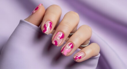 A woman's hands showcasing a trendy camouflage nail art design featuring shades of pink fuchsia and pale pink polish set against a soft lavender fabric background with subtle texture and a sheen