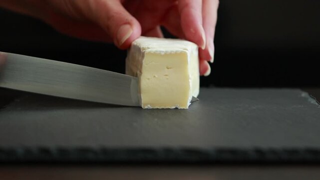 woman&rsquo;s hands carefully slicing soft brie cheese on a dark background