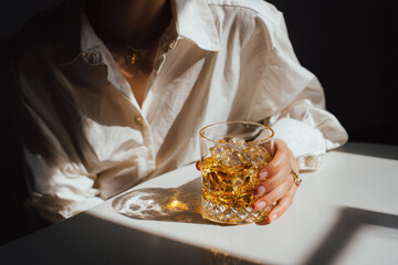 Woman hand holding glass of iced cocktail golden drink crystal glass white shirt sunlight shadow elegant refreshing close up relaxed mood
