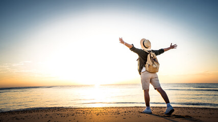 Happy man with backpack standing with arms up at the beach - Delightful tourist enjoying summer vacation by the seaside - Traveling life style and well being concept