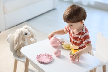 Cute little boy playing tea party with his toy bunny at table indoors
