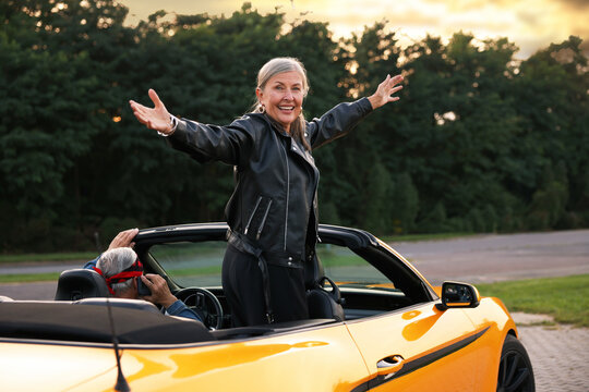 Senior couple in modern cabriolet outdoors. Woman with wide open arms and man talking on phone - Powered by Adobe