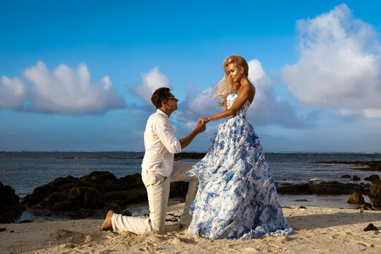Romantic couple on Mauritius tropical beach, man kneeling and kissing woman’s hand in elegant blue floral dress, love, passion and luxury fashion photoshoot at ocean sunset