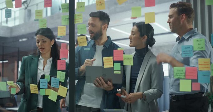 Employees Engage in a Creative Planning Session, Writing On and Placing Post-It notes on a Glass Wall to Structure a Business Project. They Discuss Each Suggestion and Rearrange Notes