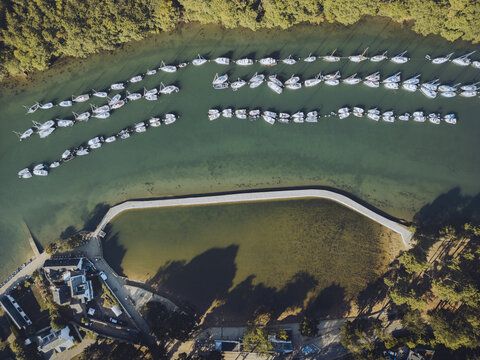 Aerial view of boats docked in the marina, reflecting the sky, with a backdrop of lush greenery and a curving walkway, Vannes, Bretagne, France.